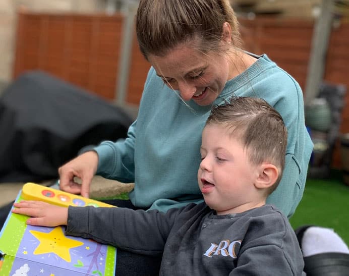 Image of carer reading a touch and feel book to a child who is living with brain tumours. The child is touching the star in the book and smiling, whilst the carer is fondly looking down at them.