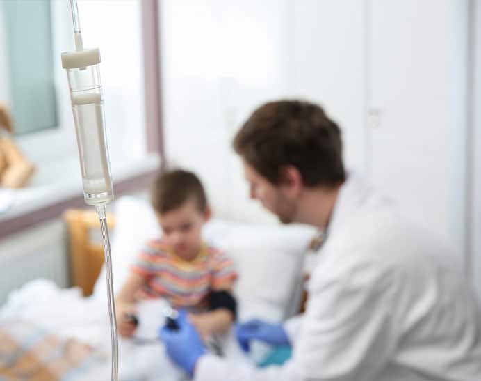 An out of focus shot of a doctor assessing a small child for the types of paediatric brain tumour they may have. There is a drip attached, and the child is sitting up whilst the doctor is taking a sample.