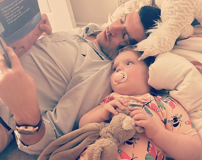 example of brain tumours in children, we have a young child laying down close to her dad who is reading her a story. She is holding her comforting toy, a brown rabbit and sucks on a pacifier.